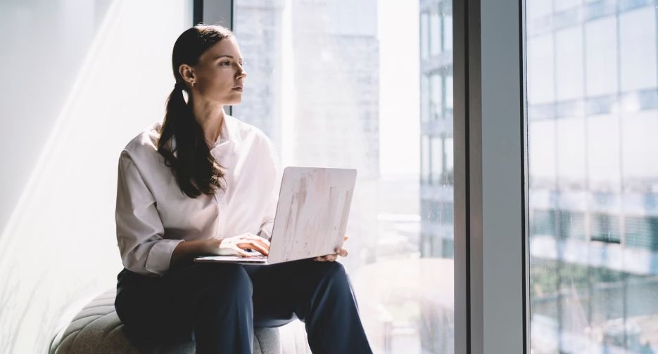 Low angle of thoughtful young female executive in casual clothes sitting on sofa and looking away while passing time at glass wall with laptop in hands
